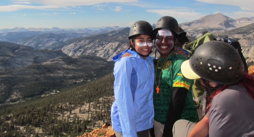 two people wearing helmets and sunscreen smile for a photo with a mountainous landscape behind them
